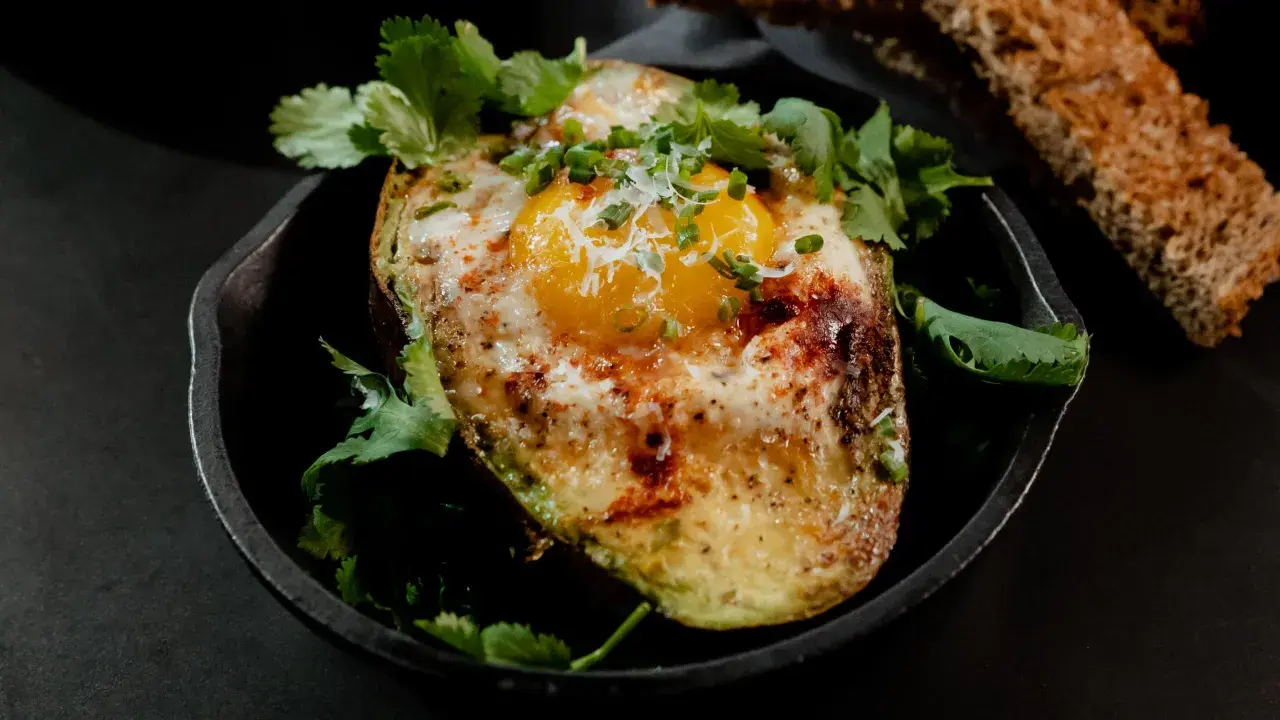 Un avocat oeuf au four, garni de coriandre fraîche et de ciboule, servi dans une poêle en fonte avec du pain grillé.
