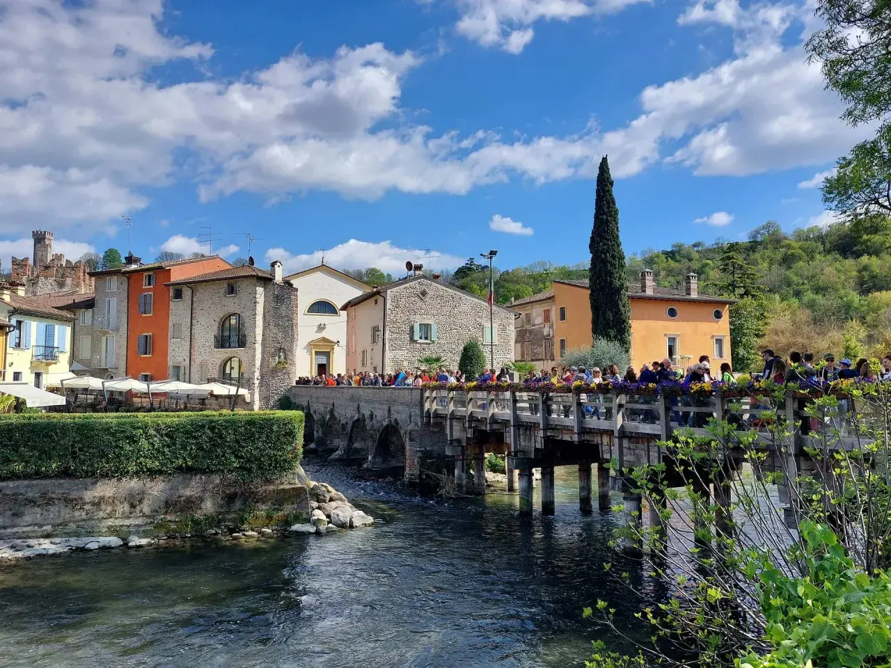 Un pittoresco borghetto mantovano con case colorate, un ponte di legno affollato di gente e un fiume che scorre sotto un cielo azzurro con nuvole bianche.