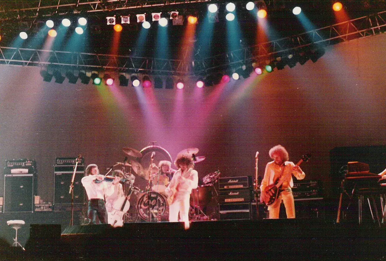 Zespół Electric Light Orchestra na scenie, grający największe przeboje. Kolorowe światła sceniczne podkreślają atmosferę koncertu.
