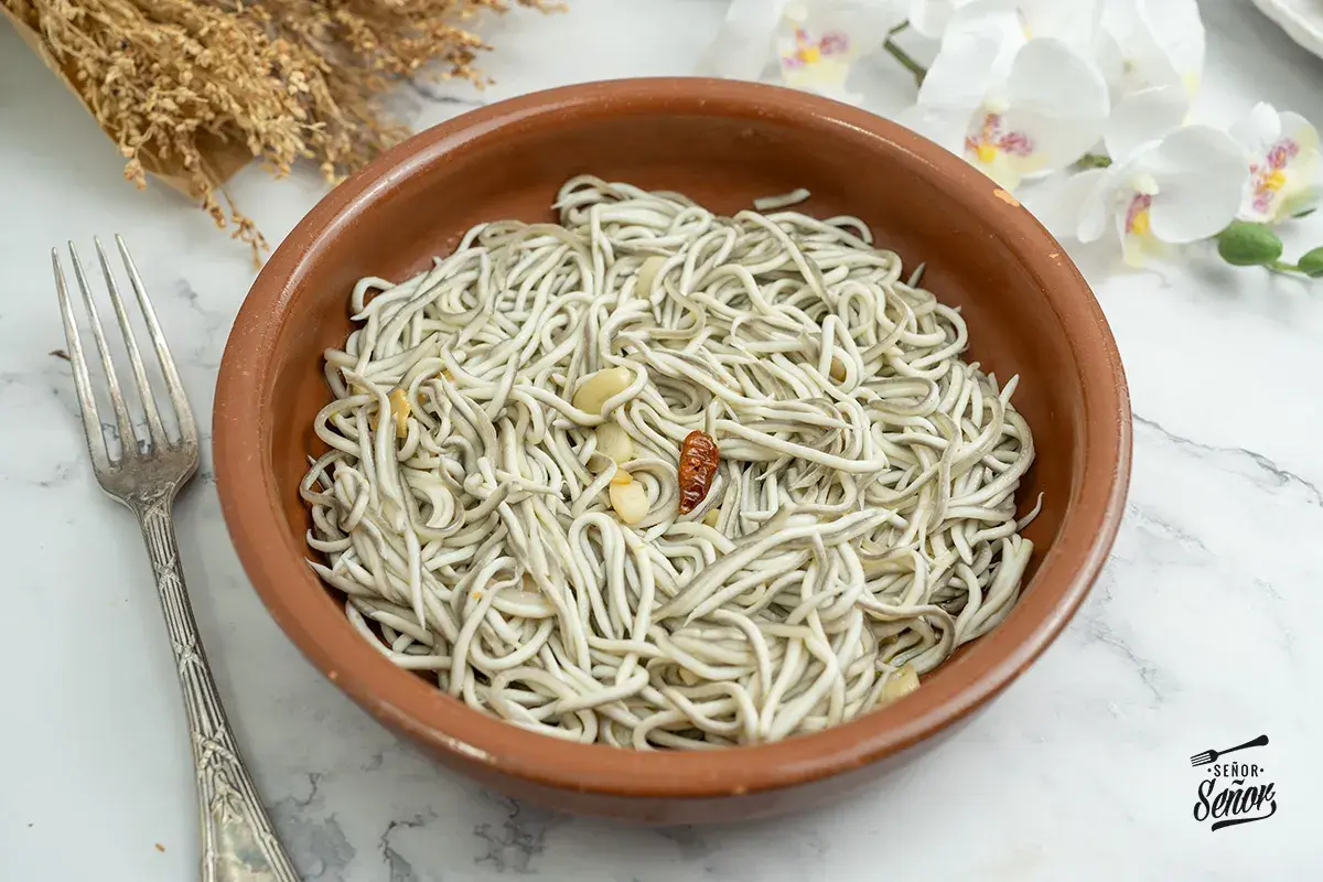 Un cuenco de barro con gulas al ajillo, ajo y guindilla. Un tenedor plateado a la izquierda y flores blancas al fondo.