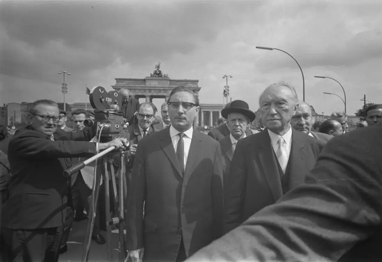 Männer in Anzügen, eine Filmkamera und das Brandenburger Tor im Hintergrund. Ein Mann hält eine "Stalin-Note" in der Hand.