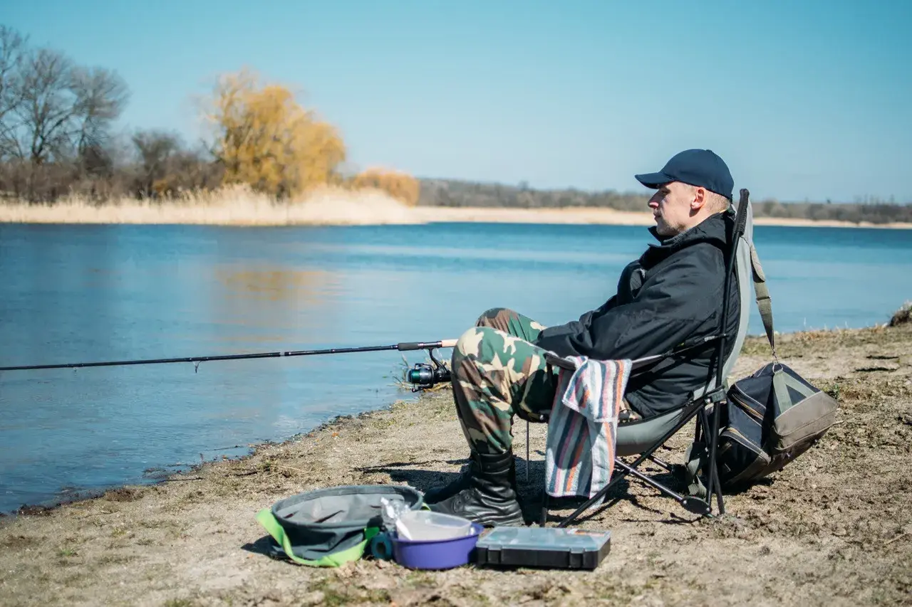 Mężczyzna siedzi na krześle nad jeziorem, łowiąc ryby metodą. Wędka zarzucona, obok torba i pudełko na akcesoria.