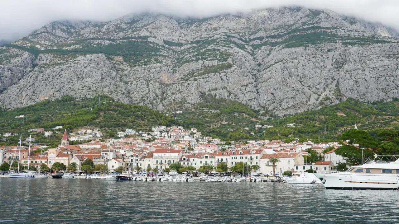 Widok na miasto Makarska z górami w tle. W porcie cumują liczne łodzie i jachty. Choć nie ma tu lotniska, widok zapiera dech w piersiach.
