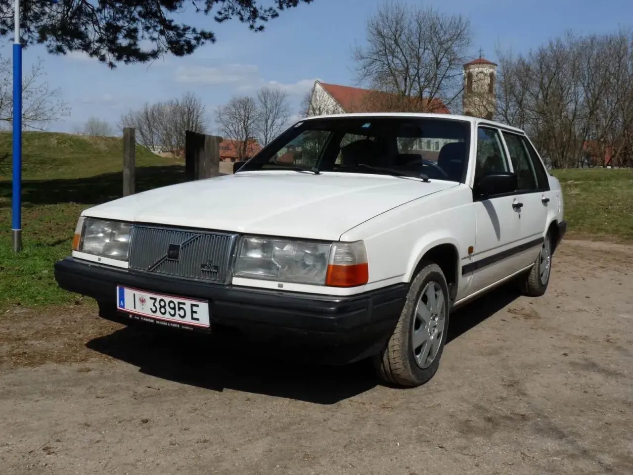Białe Volvo 840 z przodu, na tle drzew i kościoła.