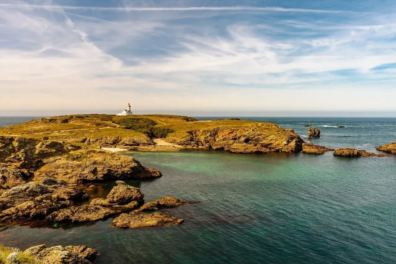 Phare blanc sur une colline verdoyante d'une île bretonne, avec des rochers dorés et une mer turquoise sous un ciel nuageux.