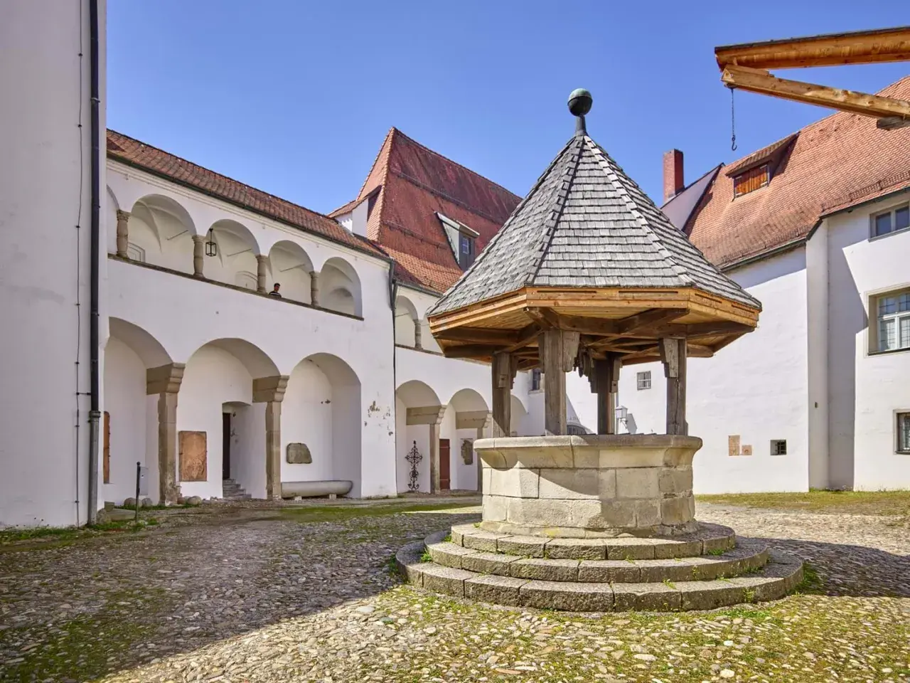 Historischer Brunnen im Innenhof des Oberhaus Passau. Weiße Arkadenbögen und rote Ziegeldächer unter blauem Himmel.