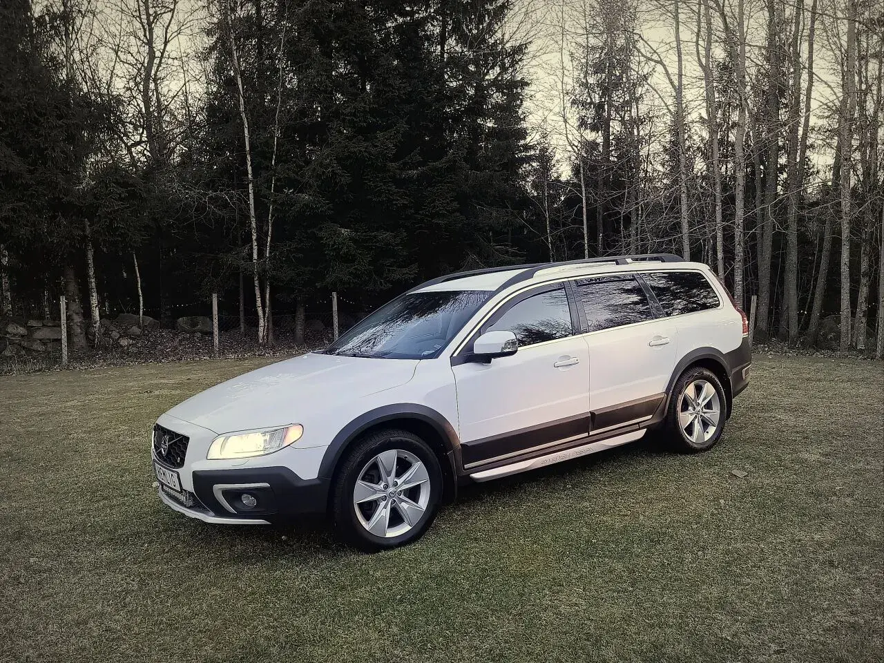 Vit Volvo XC70 parkerad på en gräsmatta med skog i bakgrunden. Kanske ett volvo d4 motor problem gör att den står stilla.