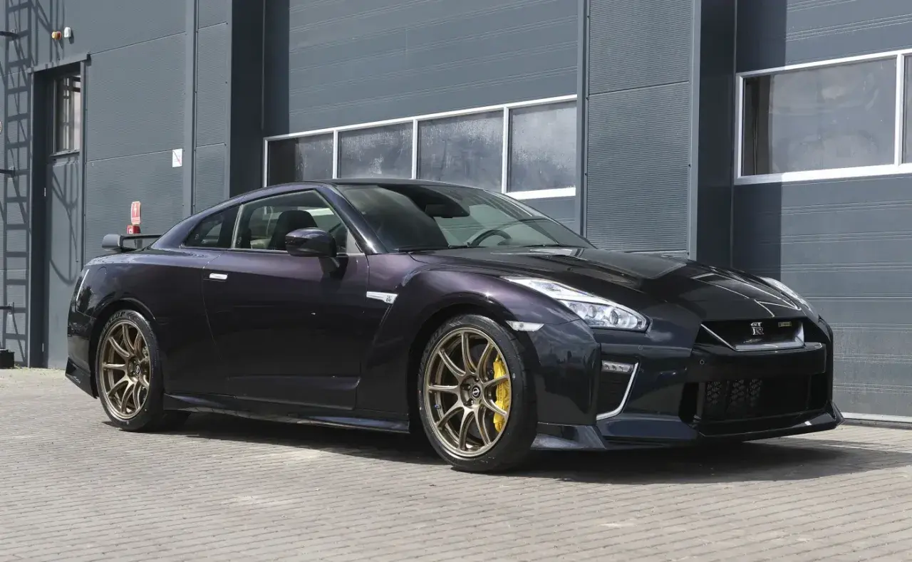 Ciemny Nissan R35 z brązowymi felgami i żółtymi zaciskami hamulcowymi stoi przed szarym budynkiem.