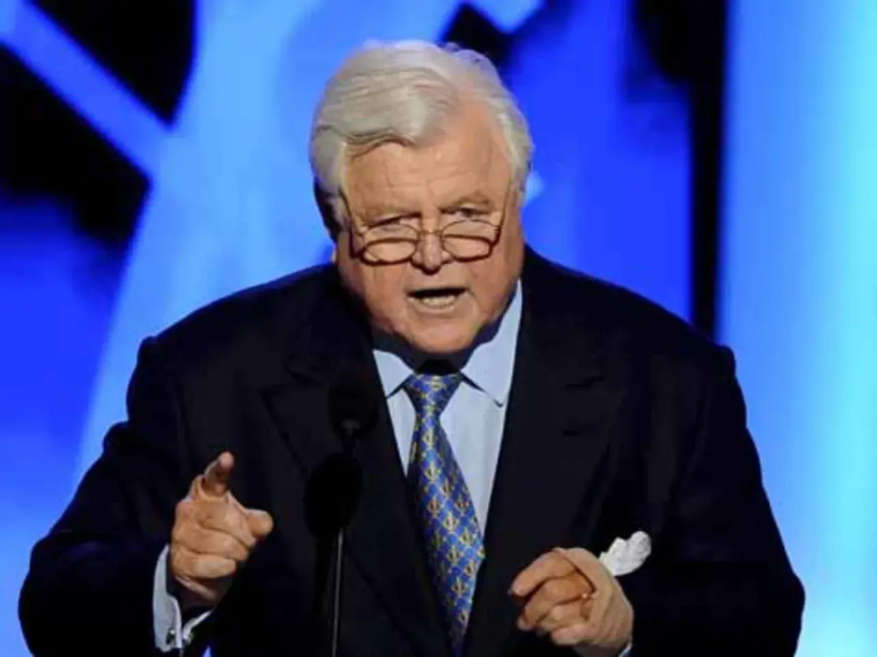 Ein älterer Mann mit Brille und grauem Haar spricht in ein Mikrofon. Er trägt einen dunklen Anzug mit einer blauen Krawatte. Seine Hand ist erhoben, als würde er eine Rede halten, die an die Kennedy-Krankheit erinnert.