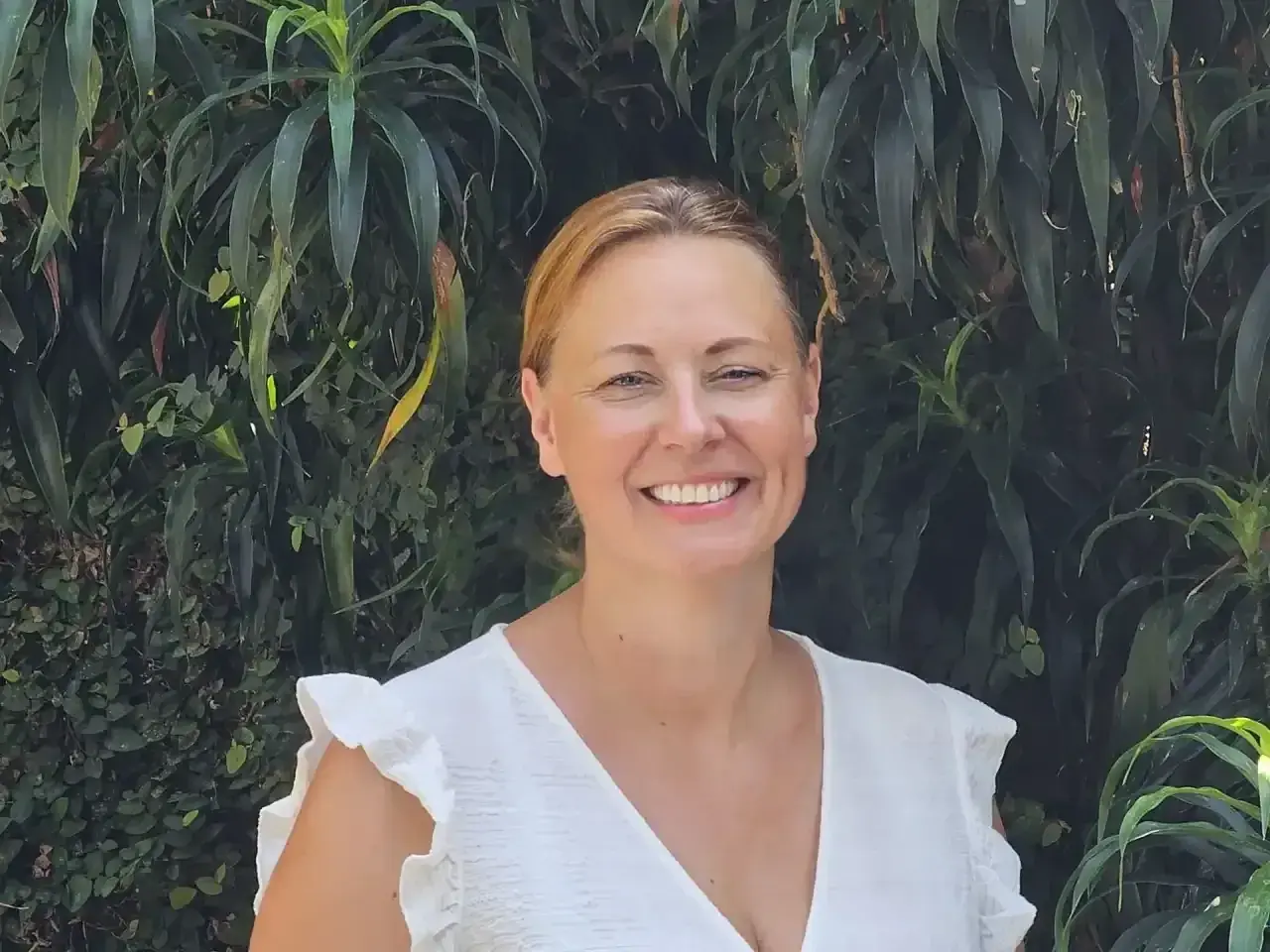 Vrolijke vrouw met blonde haren, lachend in een witte jurk, tegen een achtergrond van weelderig groen. Arjan van Dam zou trots zijn op deze foto.