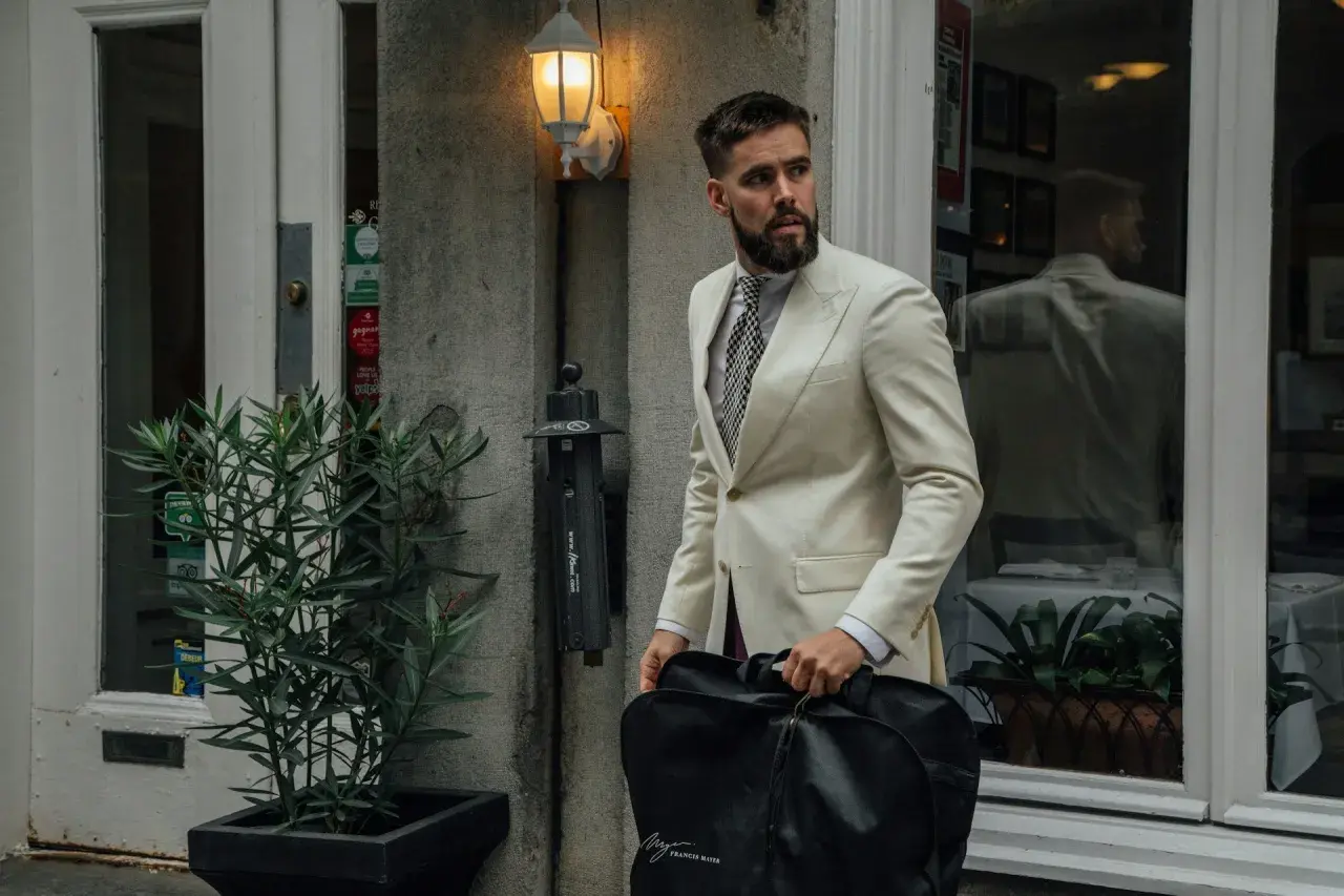 Mann im cremefarbenen Anzug mit gemustertem Hemd und Krawatte, trägt eine schwarze Kleidersacktasche. Ein lässiges Outfit für unterwegs.