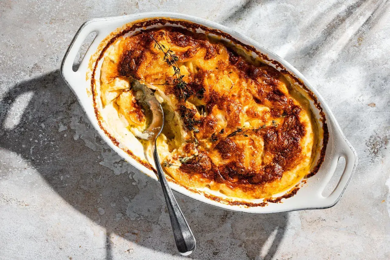 Ein goldbraun gebackenes Kartoffelgratin in einer weißen Auflaufform, garniert mit frischem Thymian. Eine silberne Löffelspitze lugt aus der cremigen Füllung hervor.
