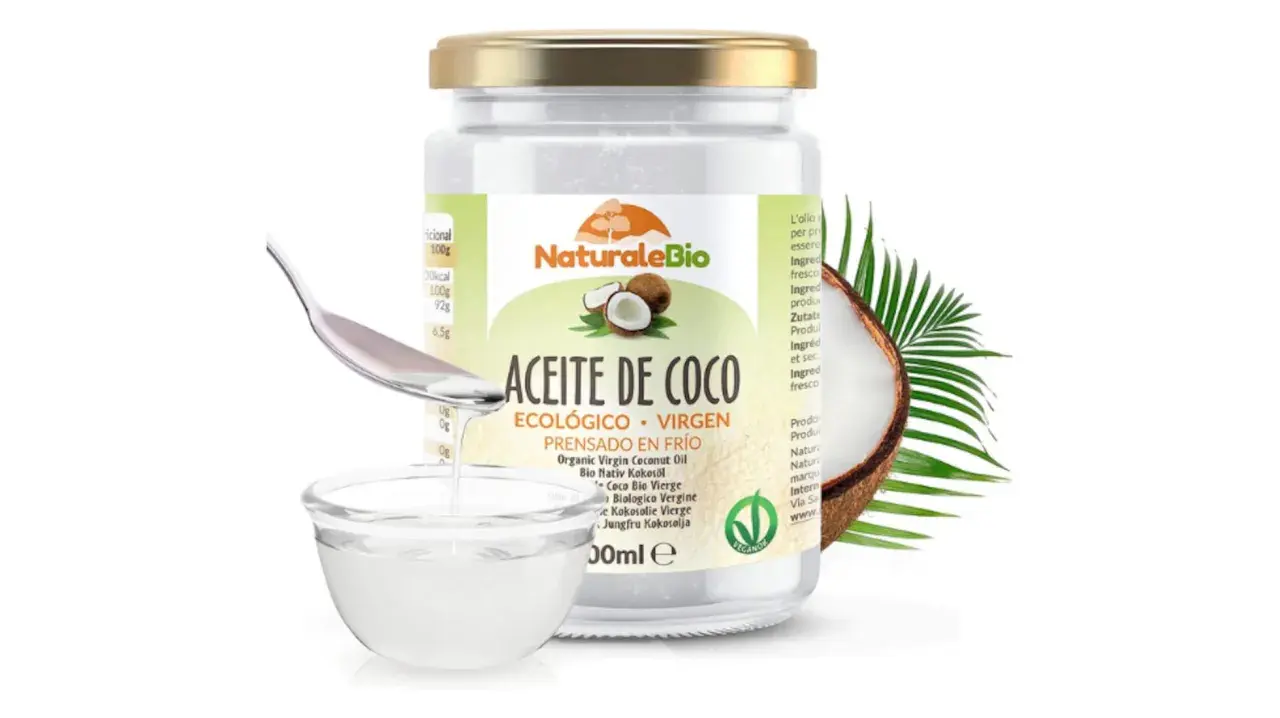 Un frasco de aceite de coco ecológico y virgen, ideal para el cuidado del pelo. Una cuchara vierte el aceite en un cuenco.