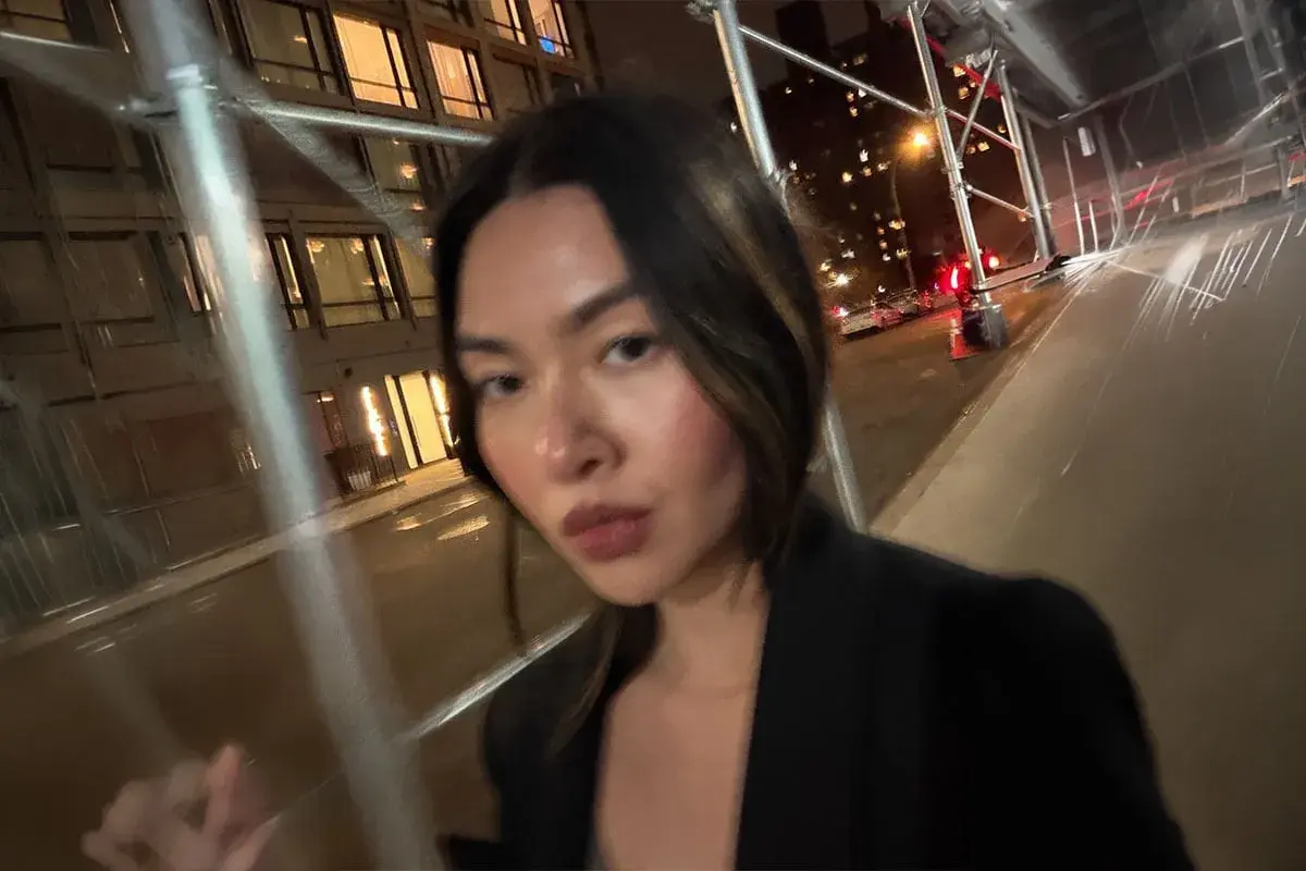 Kvinna med mörkt hår, klädd i svart kavaj, applicerar concealer utomhus på natten.