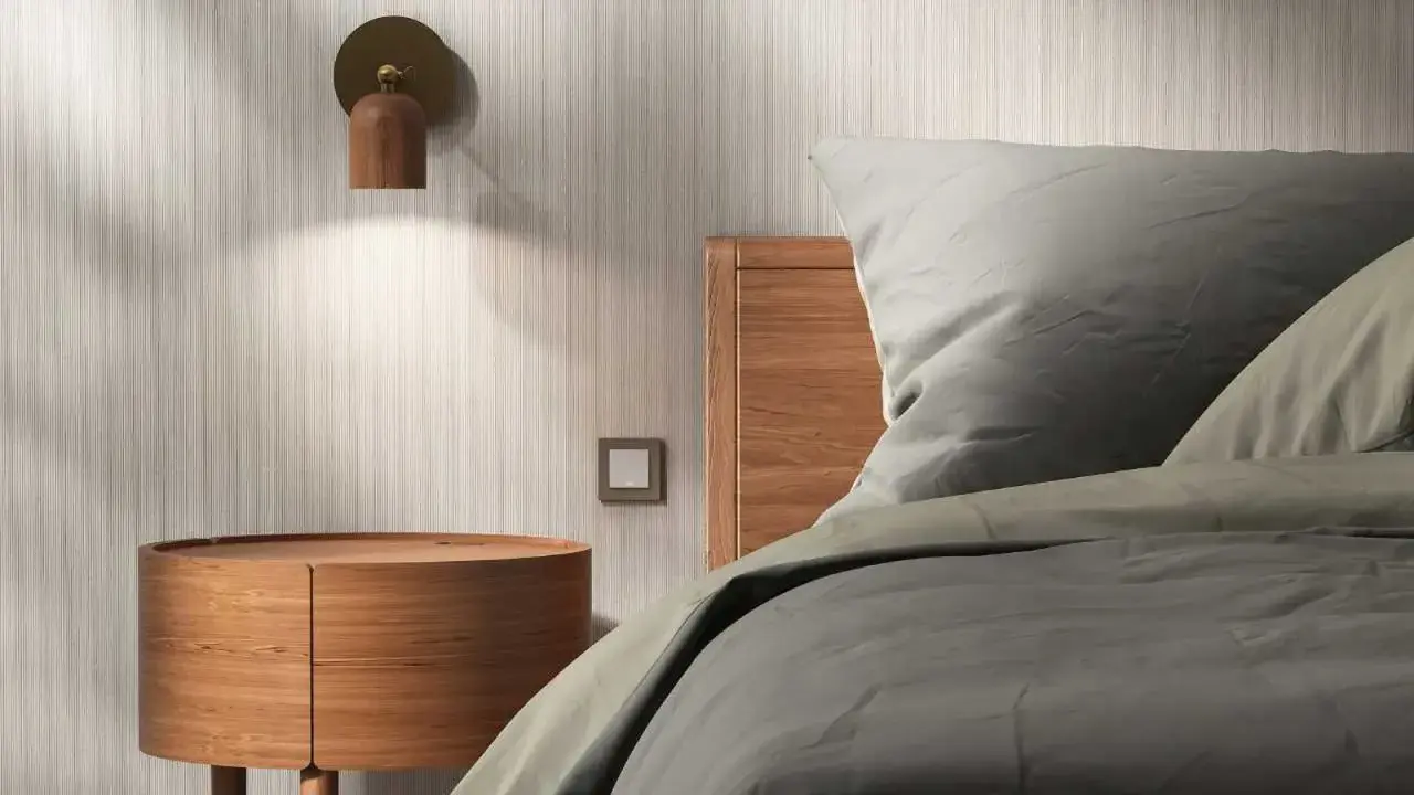 Lámpara de madera en la pared ilumina una mesita redonda y la cabecera de una cama con sábanas grises.