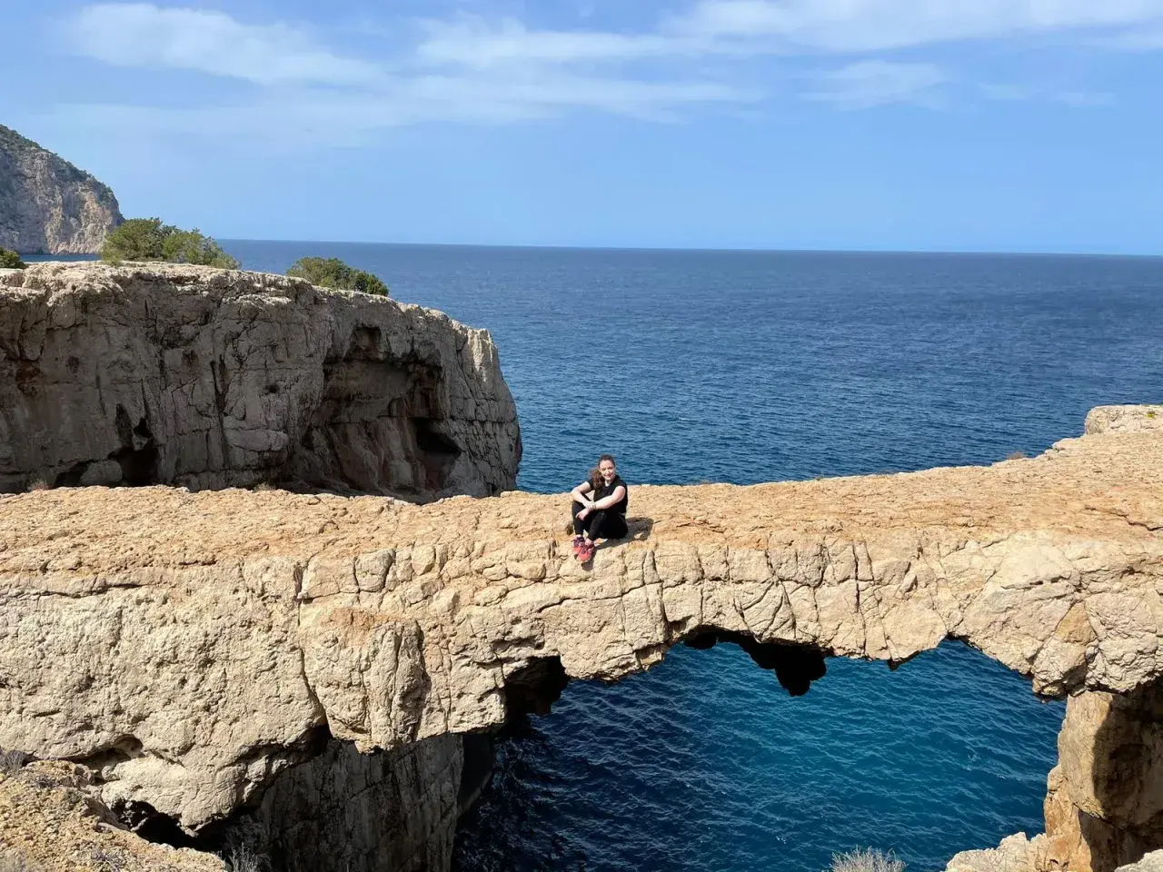 Una mujer sentada en un arco de roca natural, el "cala columpio ibiza", con el mar azul de fondo.