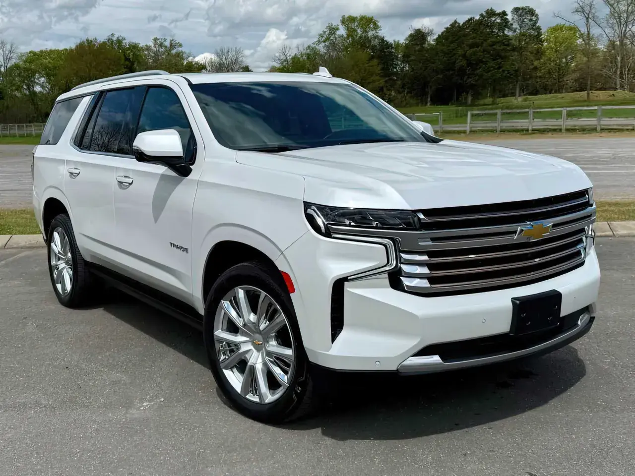 Biały Chevrolet Tahoe z chromowanymi felgami stoi na drodze, otoczony zielenią i chmurami.