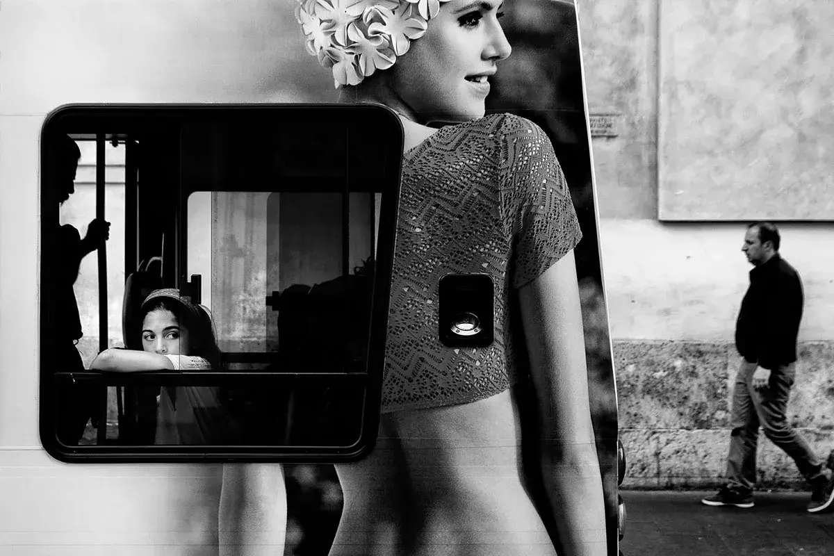 Un ritratto che evoca i grandi fotografi di moda famosi: una giovane donna con cuffia da nuoto, riflessa in un finestrino, mentre un uomo cammina sullo sfondo.