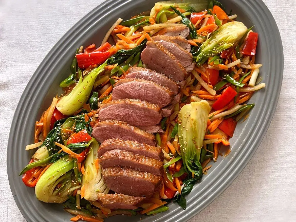 Skivad confiterad anka serveras med en färgstark wok av grönsaker som pak choi, morötter och paprika, toppad med sesamfrön.