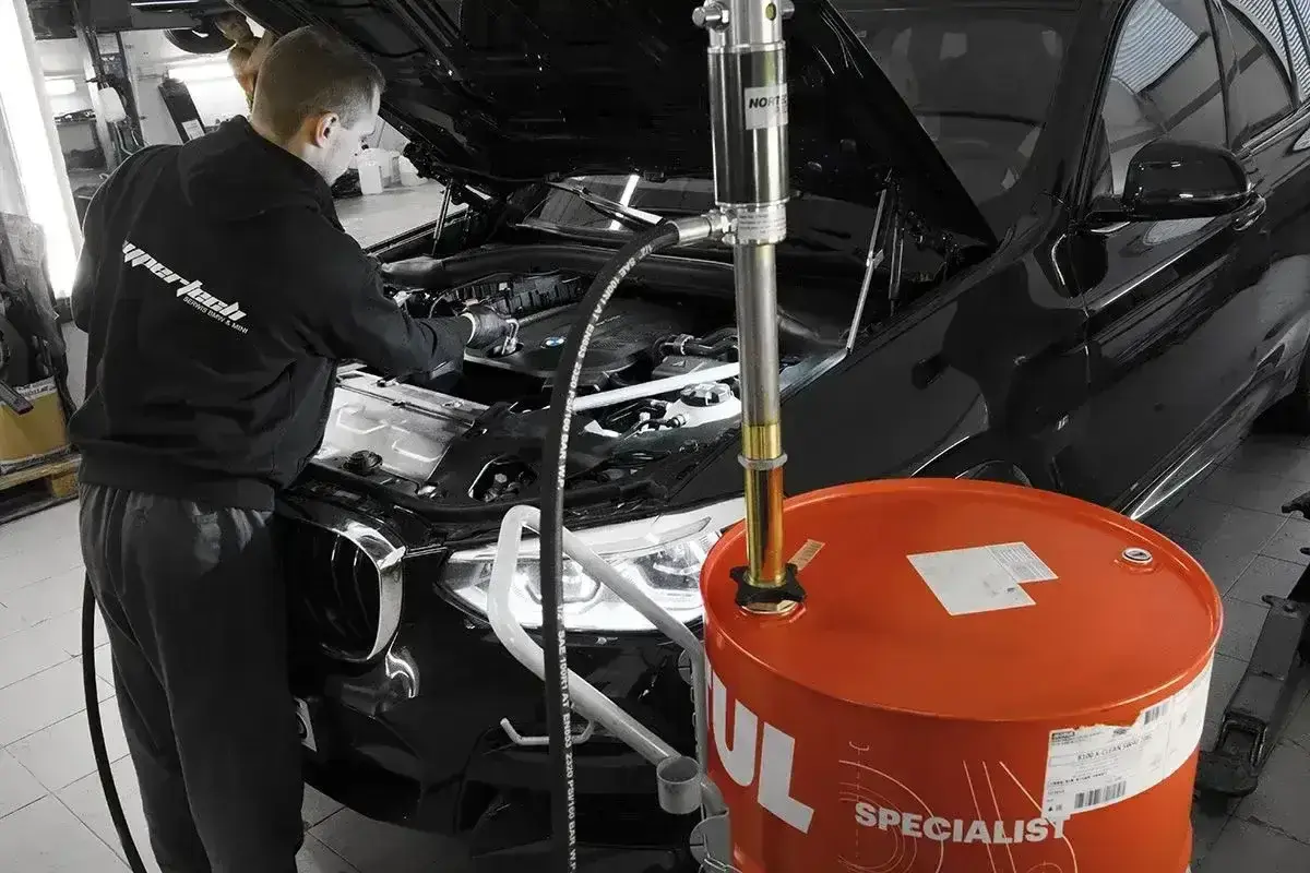 Mechanik wymienia olej w czarnym BMW. Obok stoi duża, pomarańczowa beczka z olejem. Pamiętaj, co ile wymiana oleju jest kluczowa dla silnika.