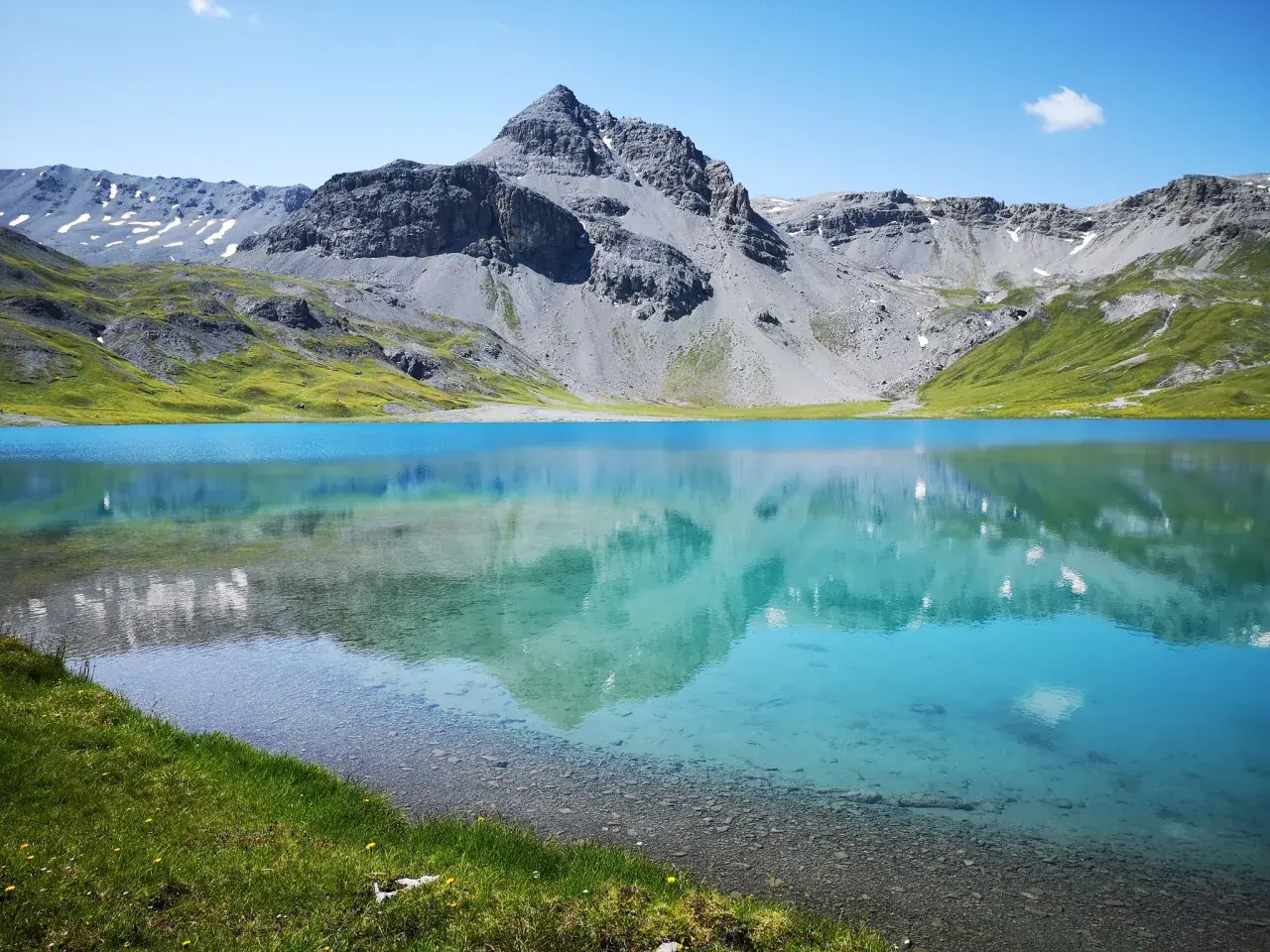 Ein klarer, blauer Bergsee, der **lai da rims**, spiegelt die majestätischen, grauen Berge und grünen Hänge wider.