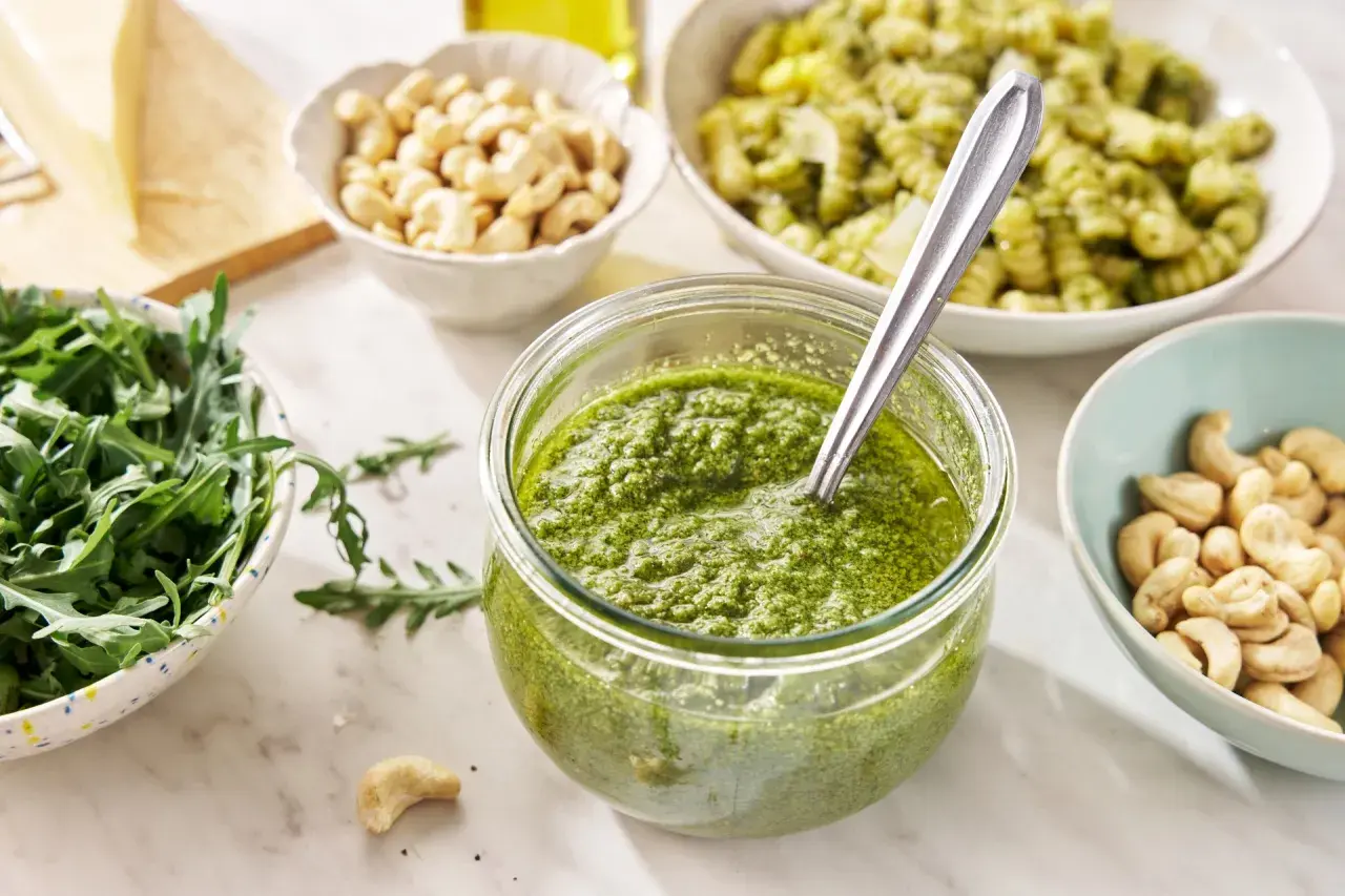 Świeże pesto z rukoli w słoiku, obok miseczki z orzechami nerkowca, rukolą i kawałkiem parmezanu.
