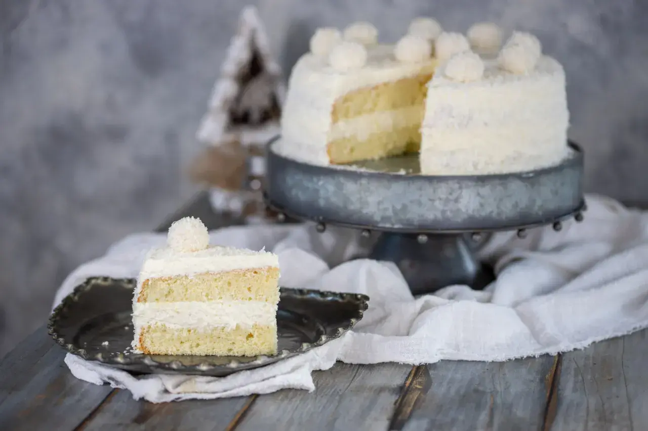 Una fetta di torta Raffaello, soffice e ricoperta di cocco, su un piatto decorato. Sullo sfondo, la torta intera su un'alzata.
