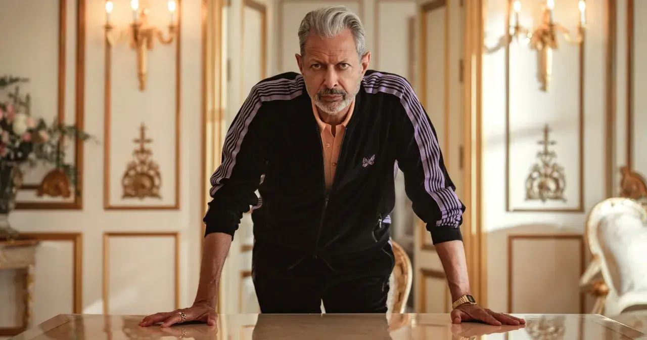 Jeff Goldblum w dresie z fioletowymi pasami, jak z jego filmów, opiera się o stół w eleganckim wnętrzu.