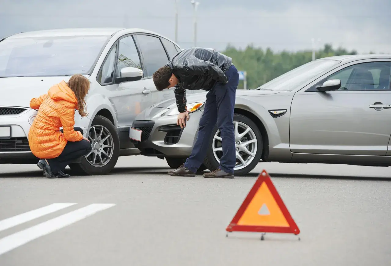 Incidente stradale con due auto danneggiate. Una donna in giacca arancione e un uomo esaminano i veicoli. Triangolo di segnalazione a terra. Nessun segno di omissione di soccorso.
