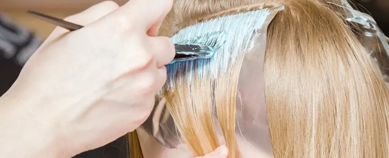 Dłoń z pędzlem nakłada jasnoniebieski rozjaśniacz na pasma blond włosów. Czas ekspozycji jest kluczowy, by uzyskać pożądany efekt.