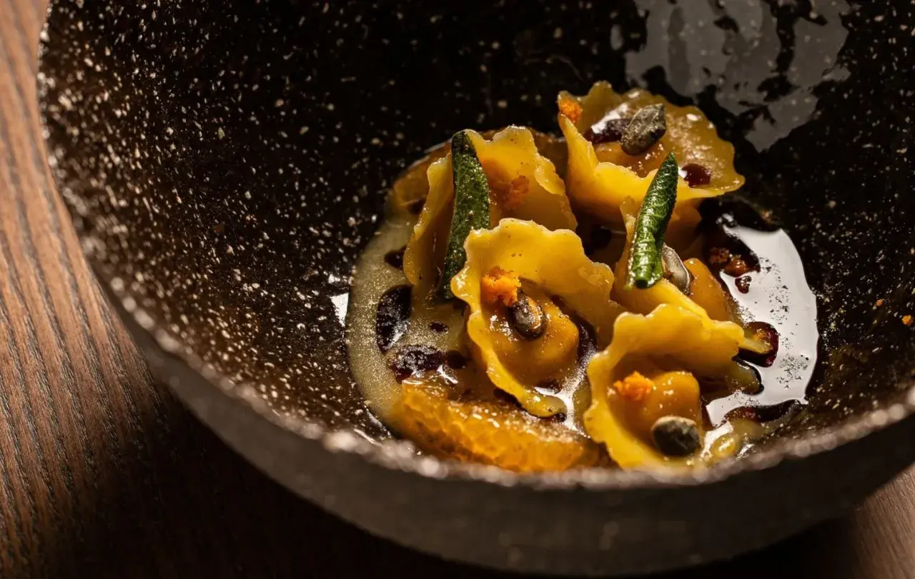 Tortelli di zucca dorati, guarniti con salvia croccante e arancia, in una ciotola scura e rustica.