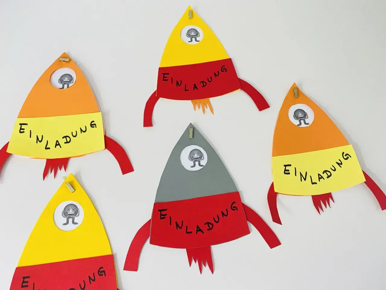 Bunte Raketen zum raketen einladung basteln, jede mit einem kleinen Monster im Fenster und dem Wort "Einladung" auf gelbem Band.