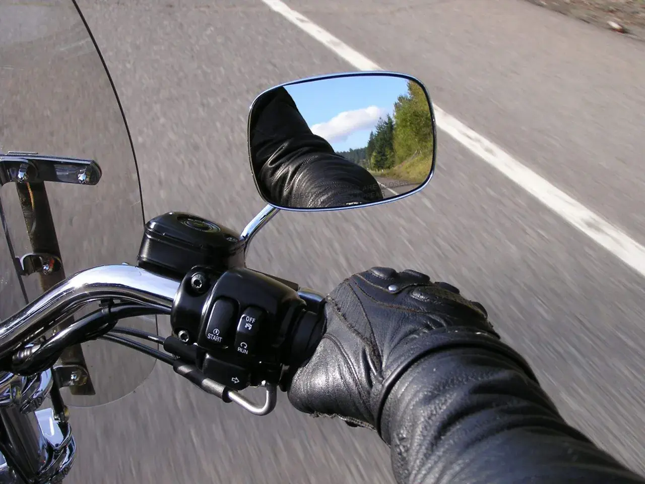 En la carretera, un motorista con guantes de cuero revisa el único espejo de su motocicleta, reflejando el cielo y los árboles.