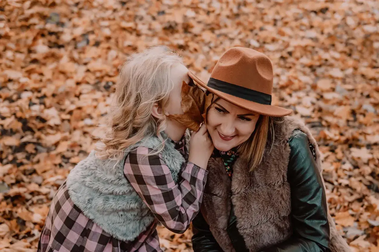 Córka szepcze mamie sekret do ucha podczas sesji mama córka wśród jesiennych liści.