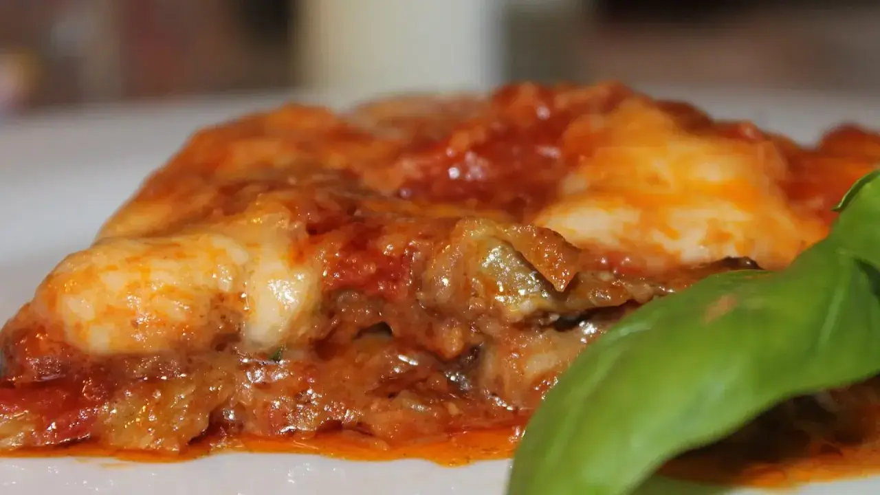 Rebanada de parmigiana di melanzane, con capas de berenjena, salsa de tomate y queso derretido, adornada con una hoja de albahaca fresca.