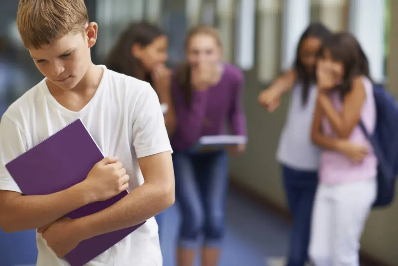 Ragazzo solo con quaderno viola, mentre altri bambini ridono e indicano. Sembra avere problemi con maestre elementari.