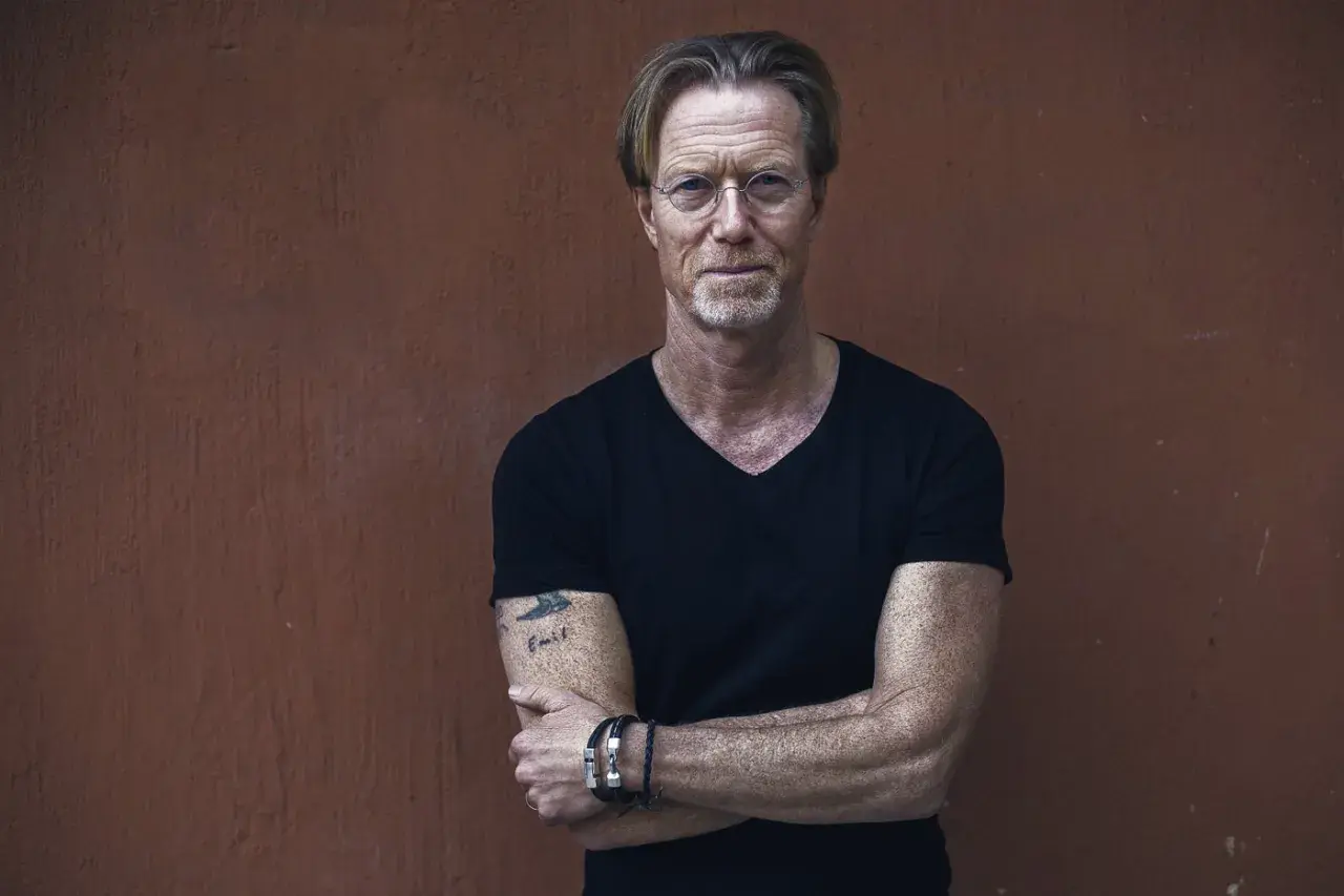 Kommissarie Ewert Grens, en man med intensiv blick och tatuering på armen, står med armarna i kors mot en rödbrun vägg.