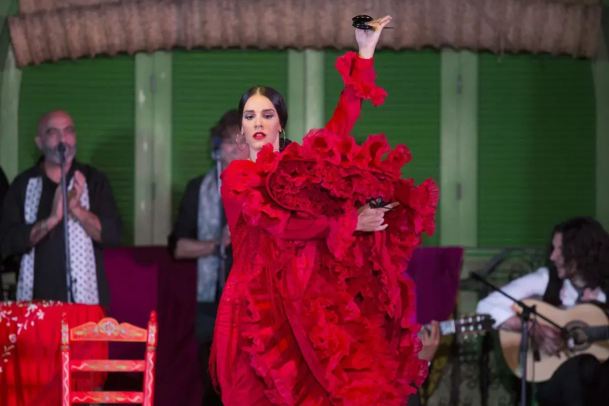 Bailaora de flamenco clásico con vestido rojo voluminoso, castañuelas en mano, acompañada por guitarrista y cantaor.