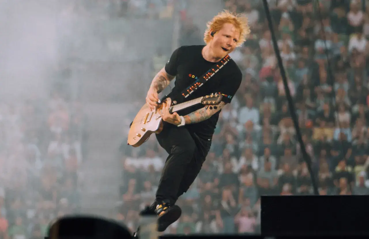 Ed Sheeran skacze na scenie, grając na gitarze, w tle tłum fanów.