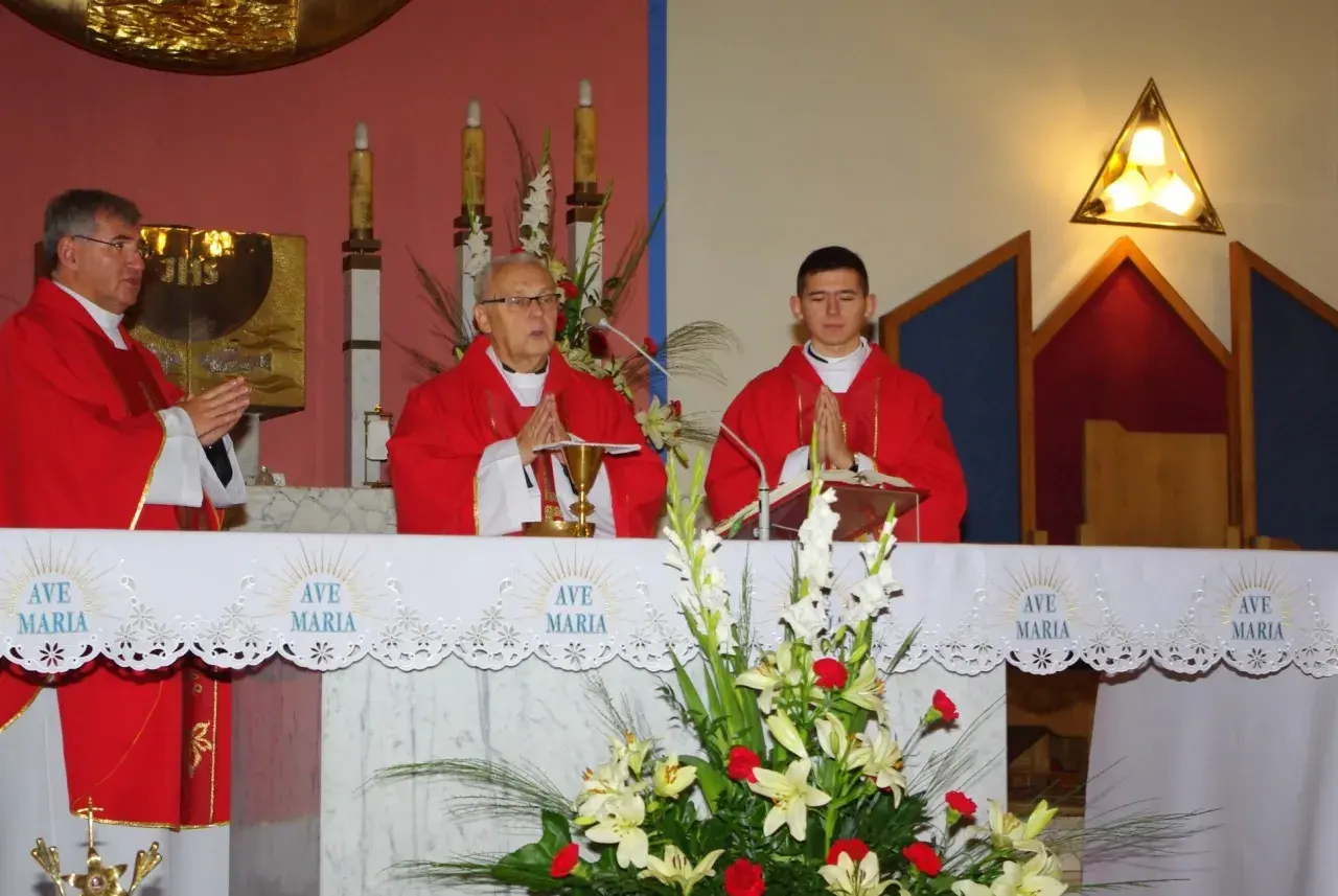 Trzech księży w czerwonych szatach podczas mszy, przed ołtarzem z kwiatami i napisem "Ave Maria".