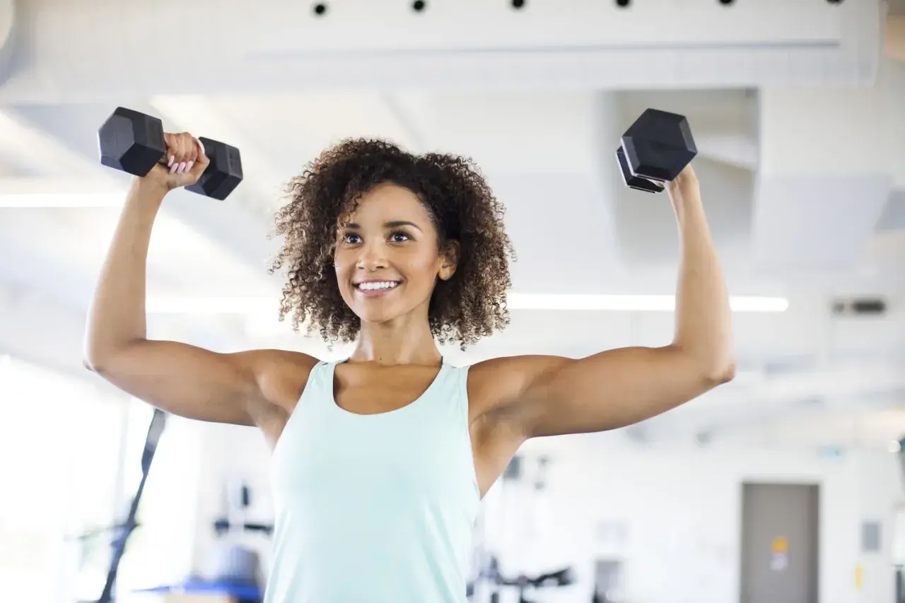 Femme souriante faisant des exercices avec haltères pour maigrir des bras. Elle est dans une salle de sport.