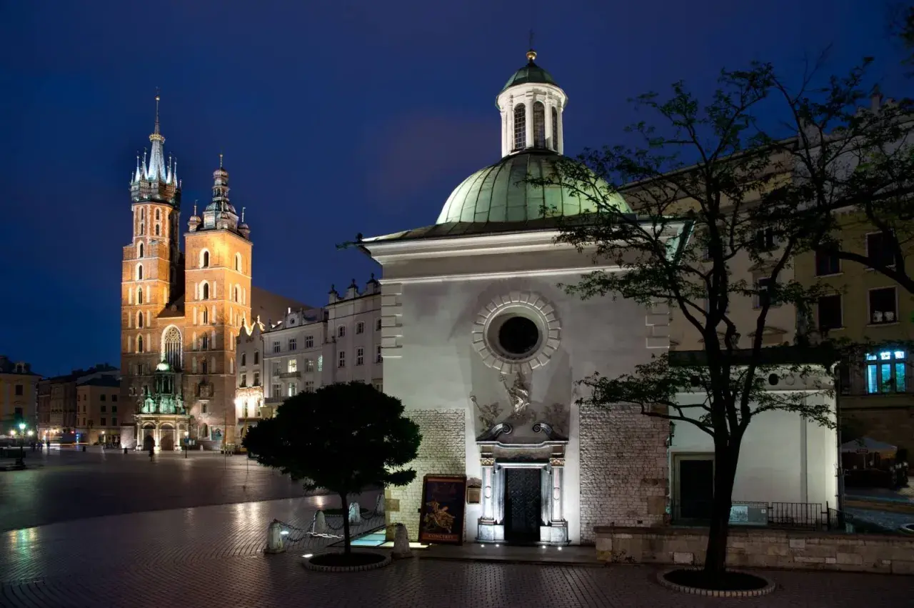 Nocny widok na Rynek Główny w Krakowie. W oddali majestatyczna Bazylika Mariacka, a na pierwszym planie kościół św. Wojciecha z zieloną kopułą.