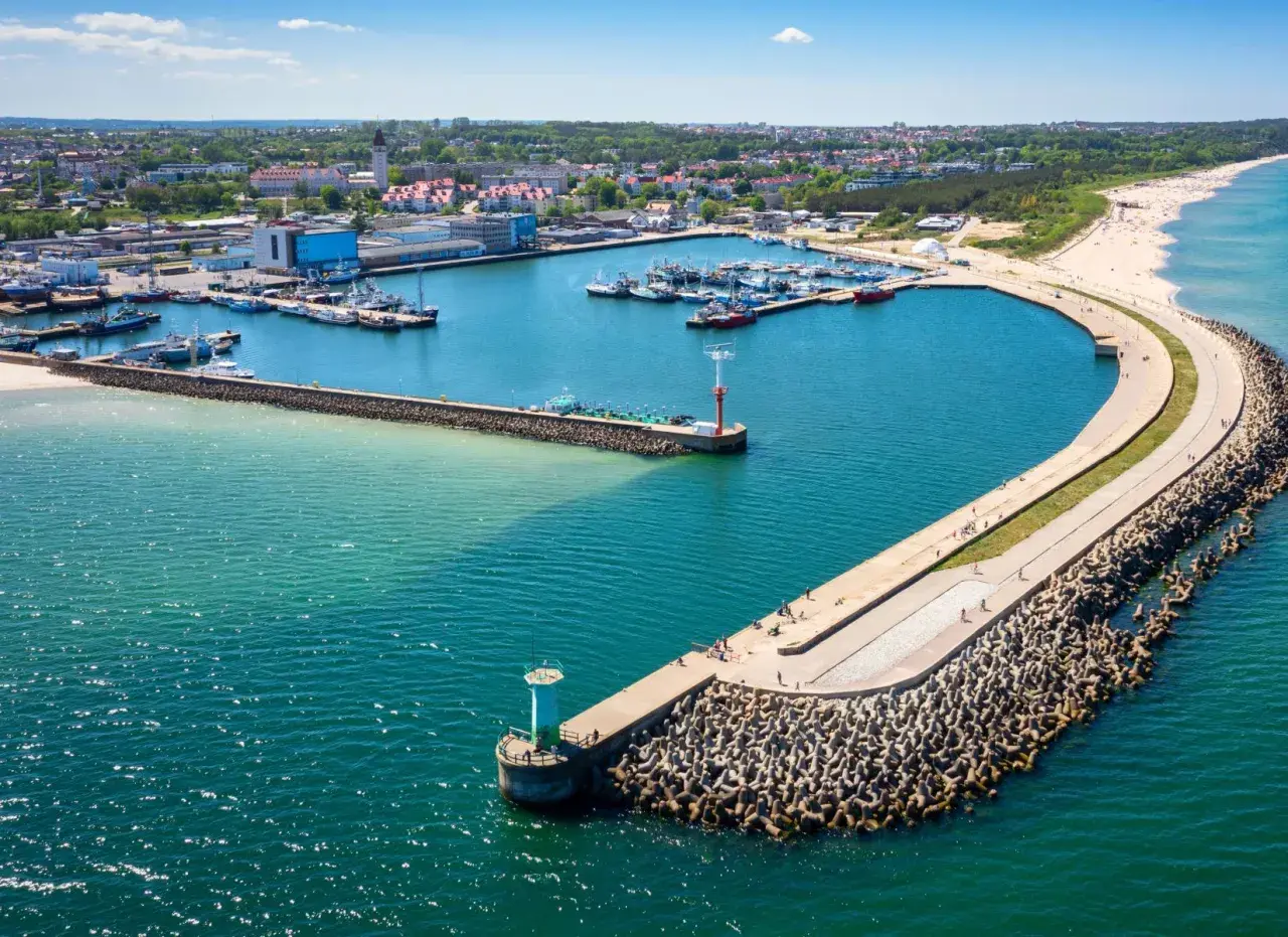 Port w Władysławowie z licznymi łodziami i falochronem, na którym widać spacerujących ludzi. W oddali plaża i zabudowania.