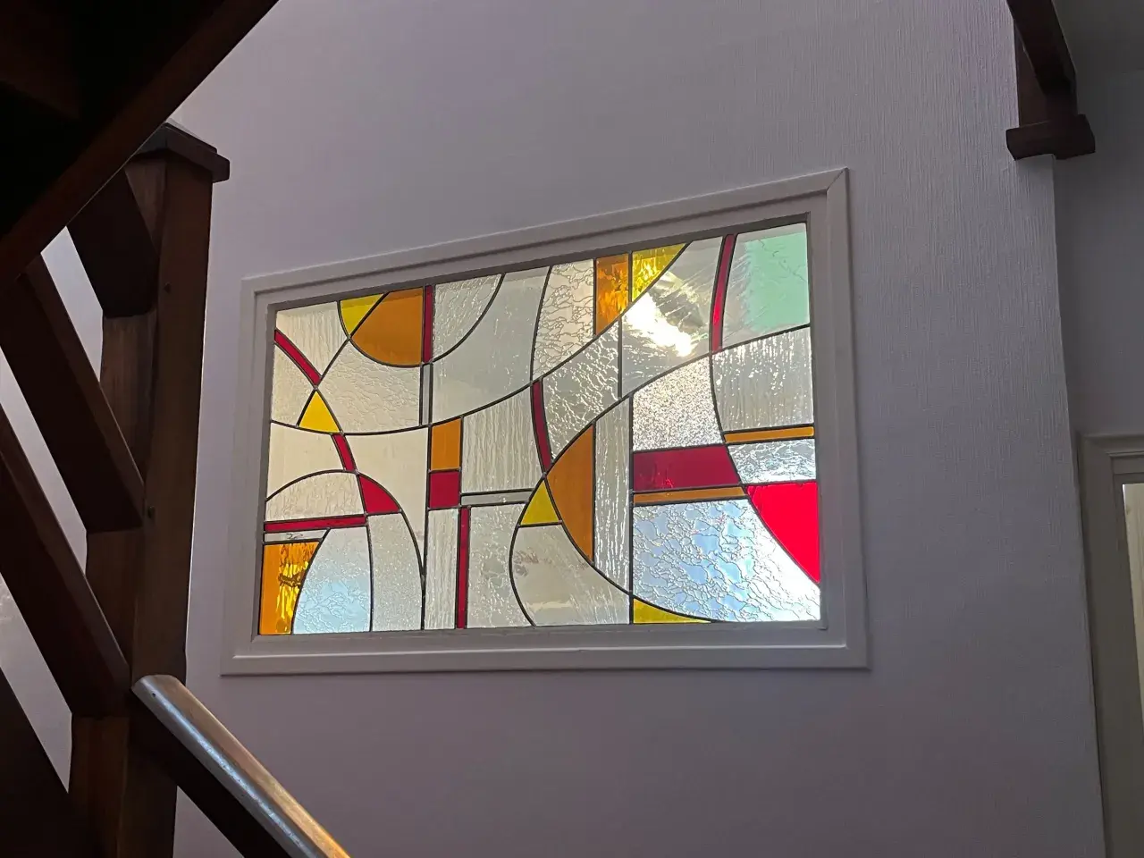 Un vitrail aux formes géométriques abstraites, aux couleurs vives et translucides, éclaire un escalier en bois. Un dessin vitrail facile à admirer.