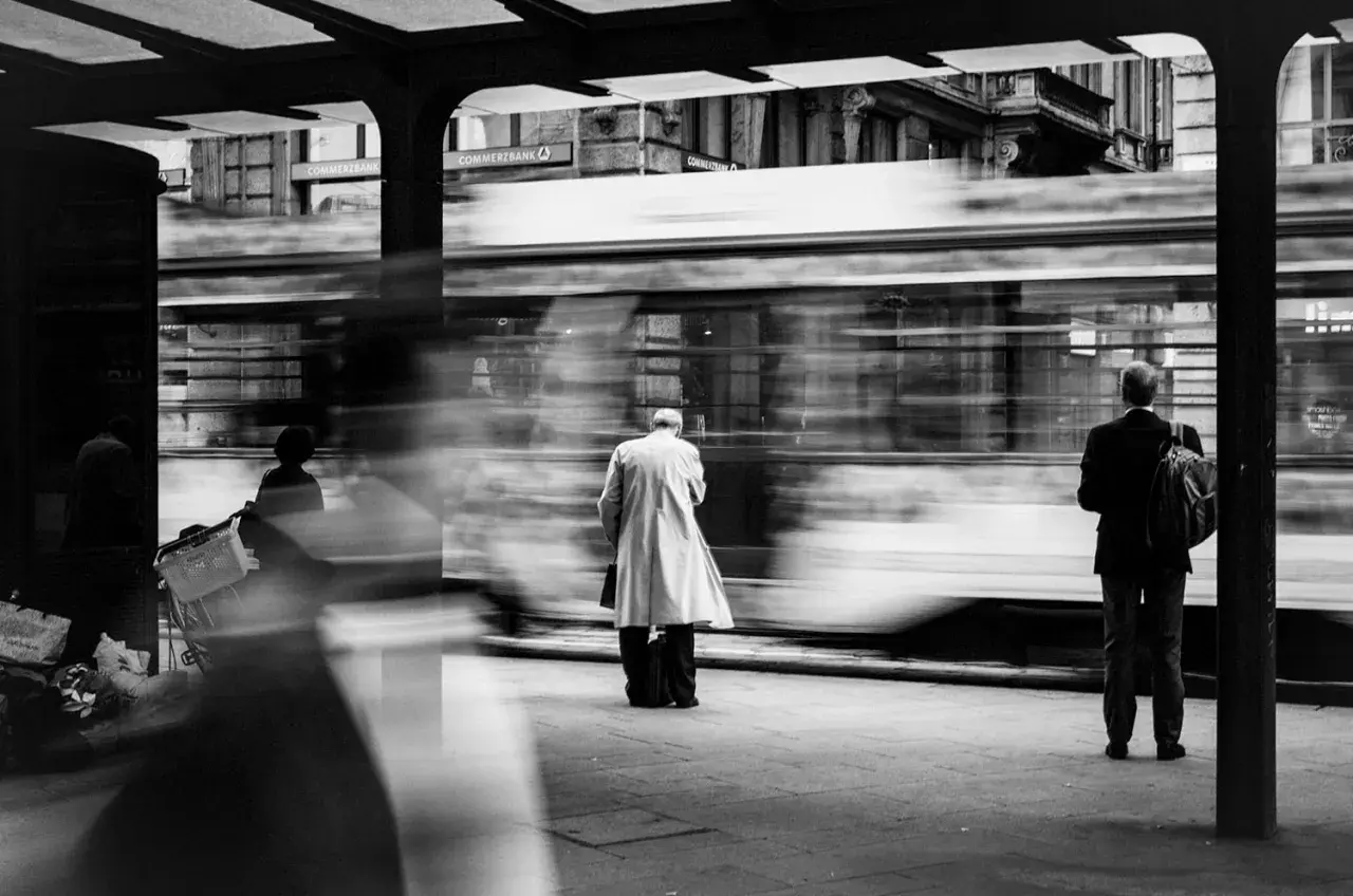 foto di strada: un uomo in impermeabile attende il tram, sfocato dal movimento. Altri passeggeri e la banca Commerzbank sullo sfondo.