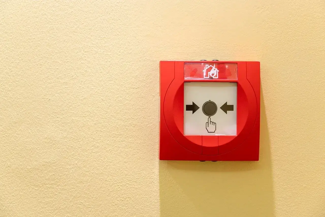 Déclencheur d'alarme incendie rouge sur mur jaune. Une main stylisée pointe le bouton, prêt à signaler une fausse alarme incendie.