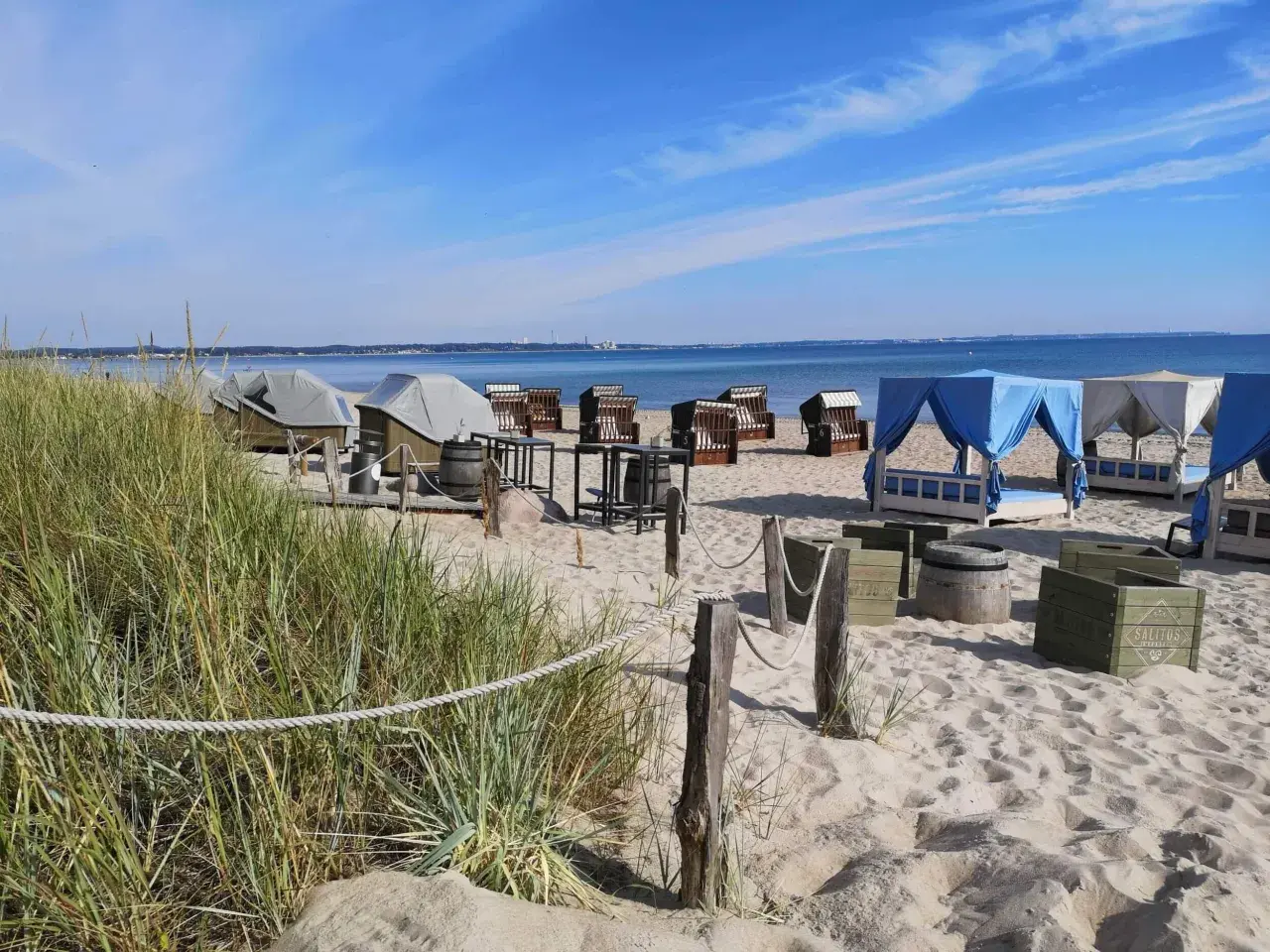 Scharbeutz Strand: Strandkörbe, Liegen unter blauen und weißen Zelten, Tische und Fässer am Sandstrand.