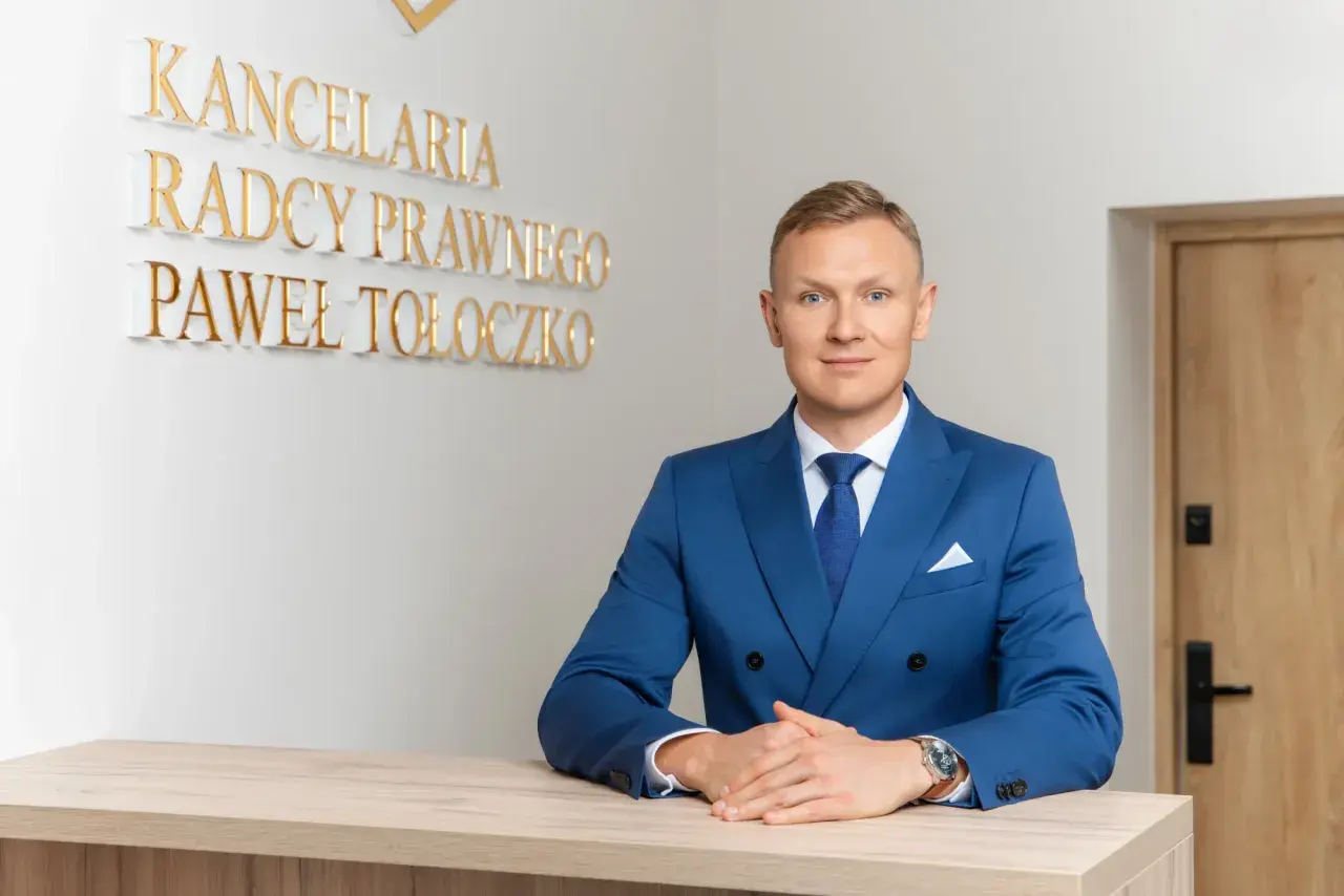 Mężczyzna w niebieskim garniturze za ladą, za nim napis "KANCELaria Radcy Prawnego Paweł Tołoczko".