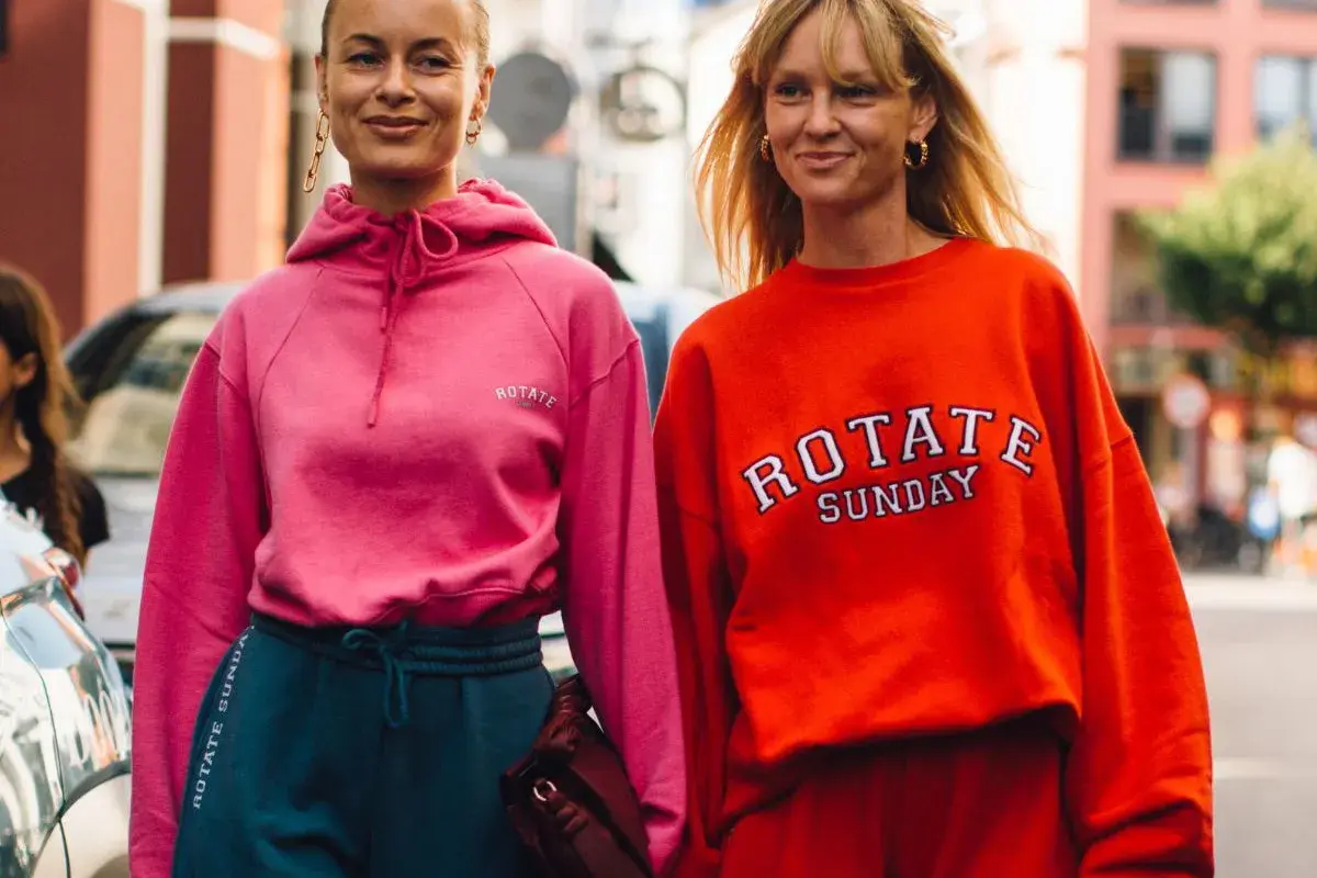 Dwie kobiety w sportowych stylizacjach, idealnych na podróż. Różowa bluza i granatowe dresy lub pomarańczowa bluza i czerwone dresy to świetny pomysł, jak ubrać się do samolotu.