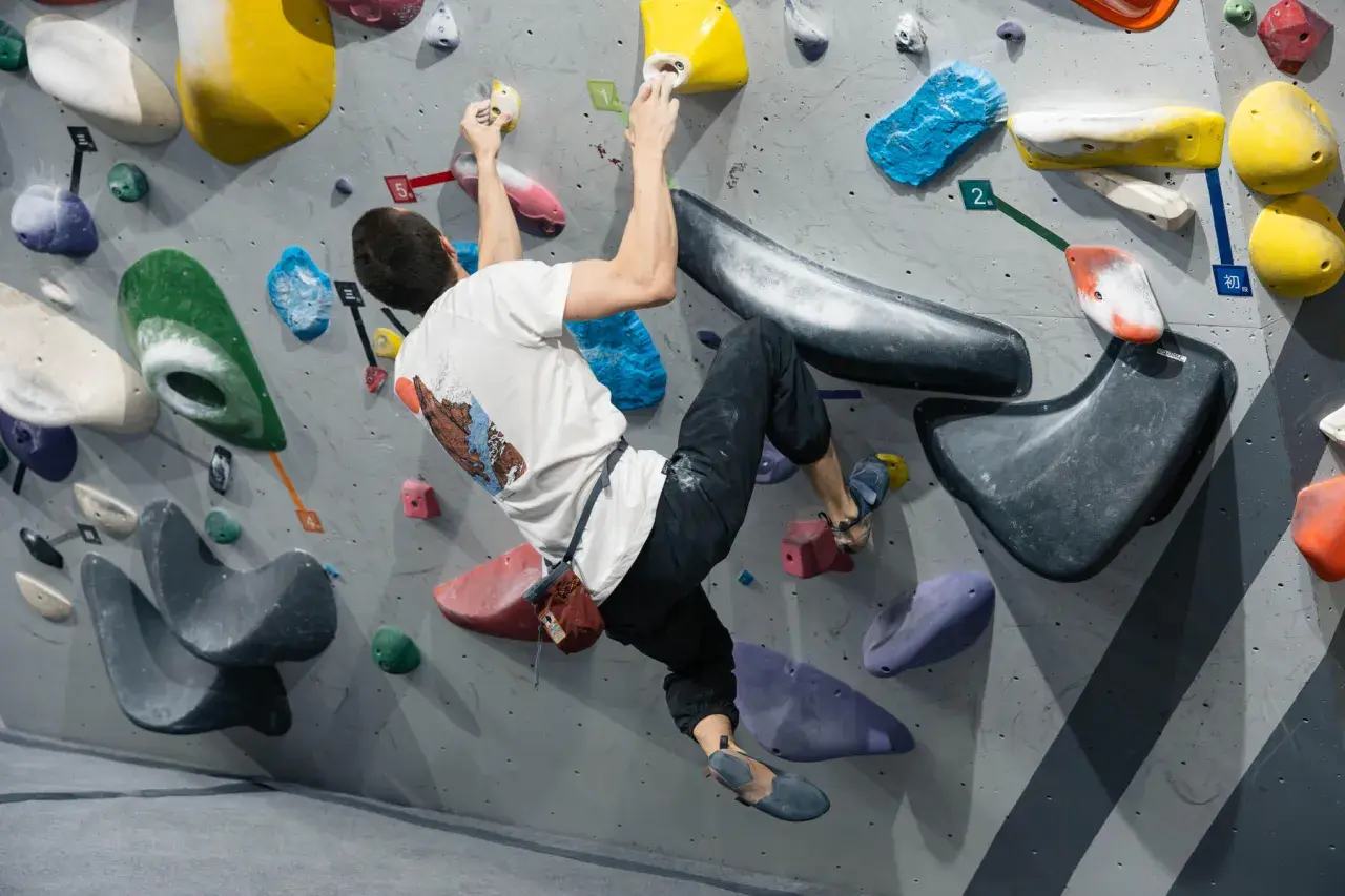Mężczyzna w białej koszulce wykonuje trudny ruch podczas wspinaczki boulderingowej na kolorowej ściance.